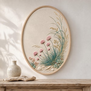May include: An oval-shaped embroidery wall art piece featuring a beige fabric background with stitched pink flowers, green grasses, and golden wheat stalks. The design includes small embroidered seashells at the base, all enclosed within a light wood frame.