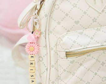May include: A white backpack with a heart pattern and a gold zipper. A pink and white keychain with the name "ERISSA" is attached to the zipper.