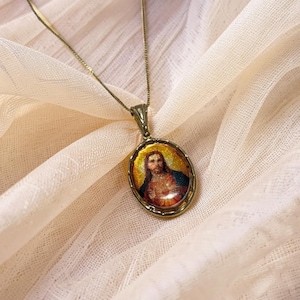 Op de afbeelding: Een goudkleurige ketting met een ovale hanger met een religieuze afbeelding. De hanger heeft een gedetailleerde illustratie met warme kleuren, omlijst door een decoratieve rand. De ketting is delicaat en de ketting rust op een zachte, crèmekleurige stof.