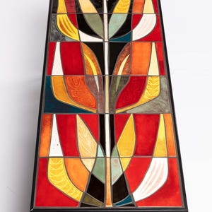May include: A rectangular coffee table with a colorful, abstract design. The tabletop features a mosaic of red, orange, yellow, black, and white geometric shapes. The table has a black metal frame and thin, angled legs.