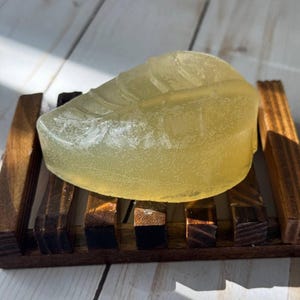 Puede incluir: Una pastilla de jabón transparente de color amarillo claro con forma de hoja, reposando sobre un jabonera de madera.