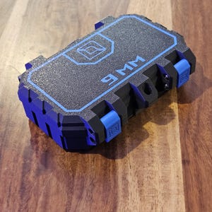 May include: A rectangular black storage case with a textured surface and blue trim. The top features a blue geometric logo and the text 9MM. The case has blue latch mechanisms on the side and a rugged, moulded exterior design with visible hinge points.