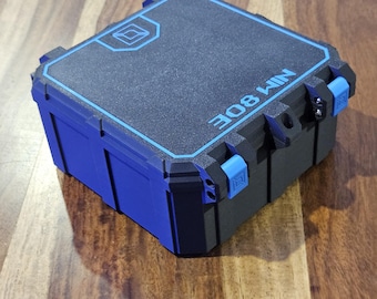 Puede incluir: Un estuche de almacenamiento cuadrado de color negro con detalles y cierres azules. La superficie superior presenta un borde azul, un pequeño logotipo geométrico y el texto "308 WIN" impreso en azul. El estuche tiene un acabado mate texturizado.