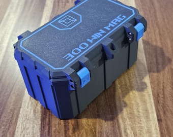 Puede incluir: Una caja de almacenamiento rectangular negra con acabado texturizado y detalles en azul. La tapa presenta un borde azul, un logotipo geométrico y el texto 300 WIN MAG. La caja incluye dos cierres azules y un bucle central para un candado.