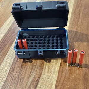 May include: A black rectangular storage case with a hinged lid and a blue interior seal. The case features a grid of circular slots designed for holding cylindrical items. Two orange shells are inside the case, and three more are placed outside.