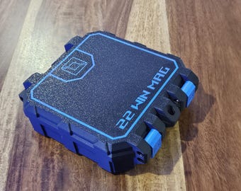 Puede incluir: Una caja de almacenamiento cuadrada de color negro y azul con una superficie superior texturizada. La caja presenta un borde azul, un logotipo geométrico y el texto 22 WIN MAG impreso en azul. Tiene cierres azules y un diseño exterior robusto.