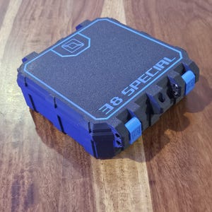 May include: A square, black storage case with blue accents and a blue border on the lid. The text 38 SPECIAL is printed in blue on the top surface, along with a small geometric logo. The case features blue latches and visible hinge pins.