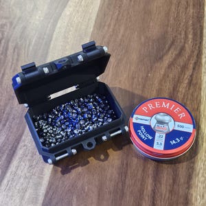Op de afbeelding: Een zwarte plastic container gevuld met .22 kaliber pellets, naast een rode, witte en blauwe blik van Premier .22 kaliber pellets. Op het blik staat "Hollow Point" en "500 count". De container is open en toont de pellets.