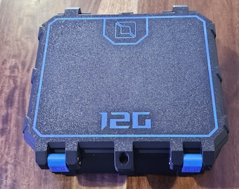 Puede incluir: Un estuche protector rígido, negro y rectangular, con una superficie texturizada. La tapa presenta un borde azul, un logotipo geométrico azul en la parte superior y el texto 12G en azul. Dos cierres azules son visibles en el borde frontal.