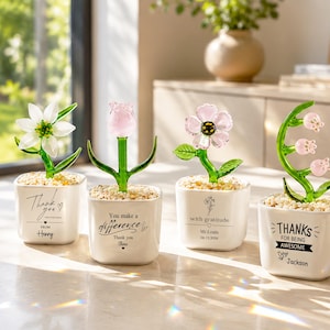 Puede incluir: Cuatro pequeñas macetas cuadradas de cerámica blanca que contienen cada una una escultura de flor de vidrio y guijarros decorativos. Las macetas presentan texto impreso en negro que incluye frases como Thank you, You make a difference, with gratitude y THANKS FOR BEING AWESOME.