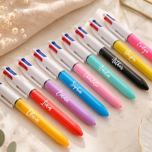 May include: An assortment of personalized pens in various colors, including yellow, red, purple, blue, pink, teal, black, and white. Each pen has a name printed on it, such as Olivia, Liam, Emma, Sophia, Charlotte, Amelia, Arthur, Harper, and Evelyn.