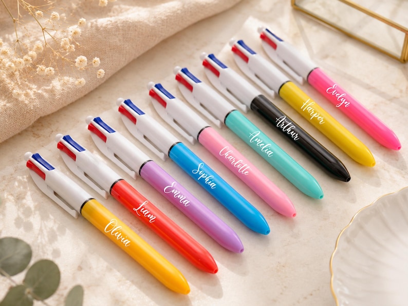 May include: An assortment of personalized pens in various colors, including yellow, red, purple, blue, pink, teal, black, and white. Each pen has a name printed on it, such as Olivia, Liam, Emma, Sophia, Charlotte, Amelia, Arthur, Harper, and Evelyn.