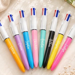 May include: Eight multi-colour pens with white barrels and ink selection buttons. Each pen has a different coloured barrel, including yellow, purple, blue, pink, teal, black, and pink. Each pen is personalised with a name in white script.