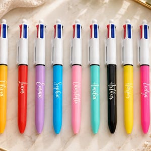 May include: A collection of ten multi-color pens with white barrels and names printed on the sides. The pens have yellow, red, purple, blue, pink, teal, black, yellow, and pink barrels. Each pen has a different name printed on it.