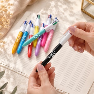 May include: A collection of colorful multi-pen writing instruments with personalized names. The pens are in various colors, including yellow, blue, green, red, purple, and pink. One black pen is held by a person, with the name "Arthur" printed on it.