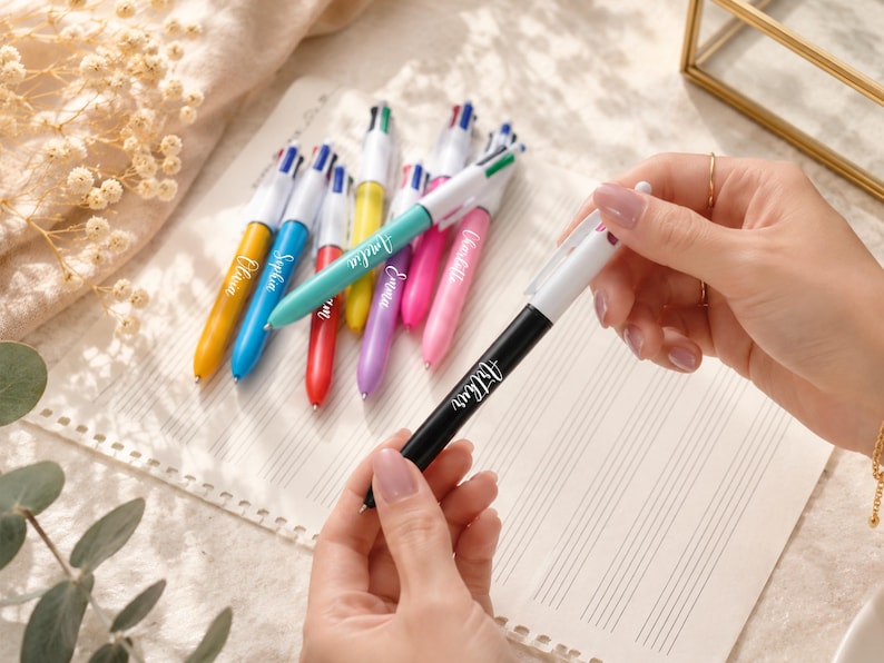 May include: A collection of colorful multi-pen writing instruments with personalized names. The pens are in various colors, including yellow, blue, green, red, purple, and pink. One black pen is held by a person, with the name "Arthur" printed on it.