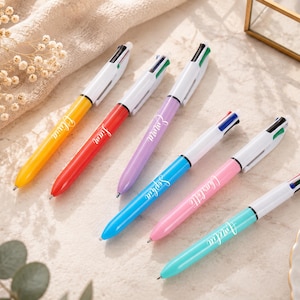 May include: Six multi-color pens with white barrels and colored tips, each with a different name in cursive script. The pens are yellow, red, purple, blue, pink, and teal. The pens are on a light-colored surface.