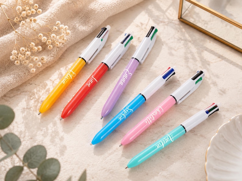May include: Six multi-color pens with white barrels and colored tips, each with a different name in cursive script. The pens are yellow, red, purple, blue, pink, and teal. The pens are on a light-colored surface.