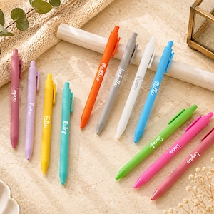 May include: A collection of vibrant pens in shades of pink, purple, yellow, green, and blue. Each pen features a name. The pens are displayed on a light-toned surface, complemented by decorative items.