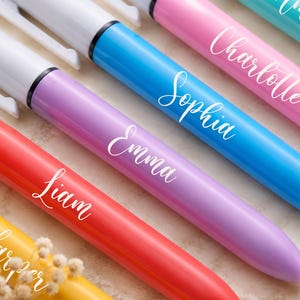 May include: A close-up of colorful personalized pens. The pens are in shades of yellow, red, purple, blue, and pink. Each pen has a white name printed on it in a cursive font. The names include Harper, Liam, Emma, Sophia, Charlotte, and Amelia.