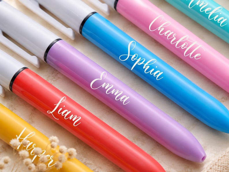 May include: A close-up of colorful personalized pens. The pens are in shades of yellow, red, purple, blue, and pink. Each pen has a white name printed on it in a cursive font. The names include Harper, Liam, Emma, Sophia, Charlotte, and Amelia.