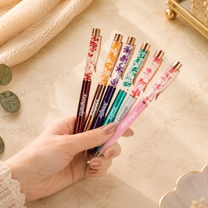 Puede incluir: Una mano sosteniendo cinco bolígrafos coloridos con diseños florales. Los bolígrafos tienen barriles de diferentes colores, incluyendo rojo, morado, turquesa y rosa. Cada bolígrafo tiene una punta dorada y un nombre impreso en el lateral.