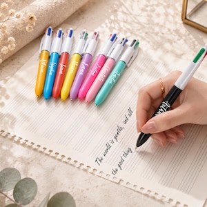 May include: A collection of colorful ballpoint pens with white barrels and multi-colored ink tips. Each pen has a different name printed on the barrel. A hand is writing on lined paper with a black and white pen. The text on the paper reads "The world is gentle, and all the good thing".