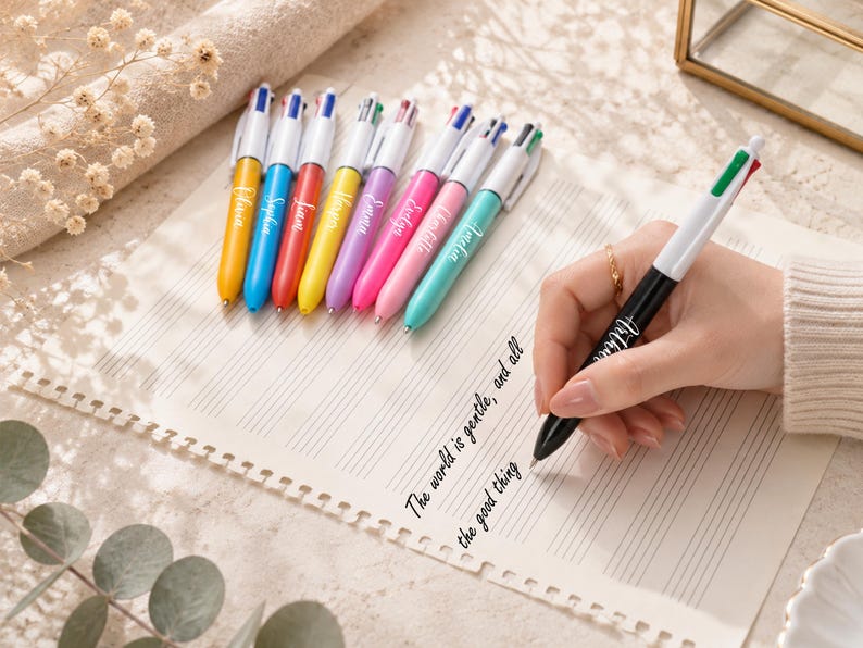 May include: A collection of colorful ballpoint pens with white barrels and multi-colored ink tips. Each pen has a different name printed on the barrel. A hand is writing on lined paper with a black and white pen. The text on the paper reads "The world is gentle, and all the good thing".