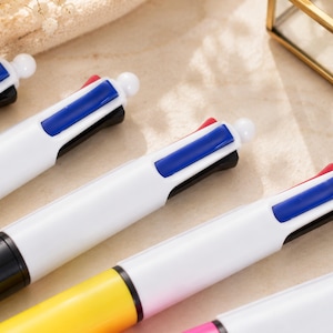 May include: Close-up of several multi-color pens. Each pen has a white barrel, a black tip, and a button with red, blue, and black ink options. The pens are arranged diagonally, with the tips facing the lower right corner.
