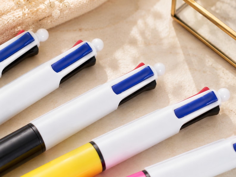 May include: Close-up of several multi-color pens. Each pen has a white barrel, a black tip, and a button with red, blue, and black ink options. The pens are arranged diagonally, with the tips facing the lower right corner.