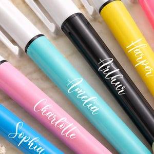 May include: Close-up of several colorful pens with white ink. The pens are in shades of blue, pink, yellow, and black. Each pen has a name written on it in a cursive font: Sophia, Charlotte, Amelia, Arthur, Harper, and Evelyn.