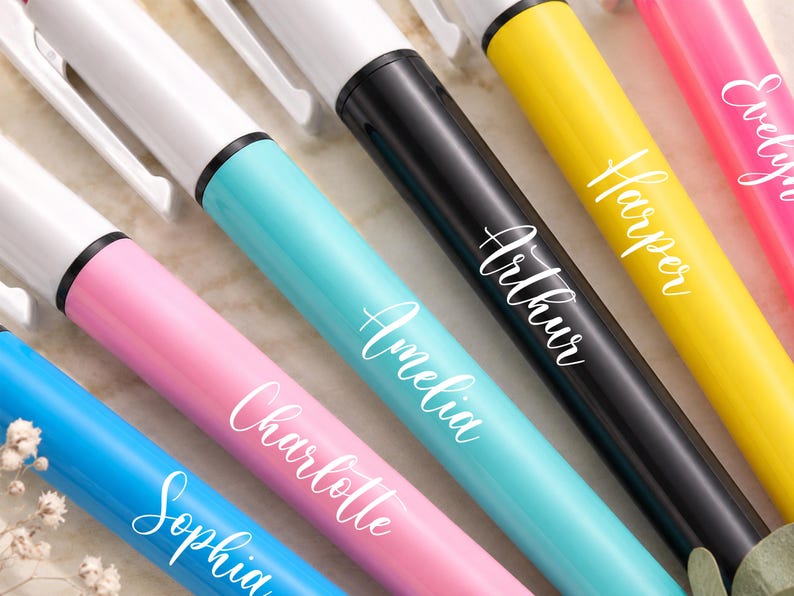 May include: Close-up of several colorful pens with white ink. The pens are in shades of blue, pink, yellow, and black. Each pen has a name written on it in a cursive font: Sophia, Charlotte, Amelia, Arthur, Harper, and Evelyn.