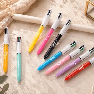 May include: A collection of colorful multi-pen ballpoint pens. Each pen has a white barrel and a different colored body, including yellow, pink, black, blue, pink, purple, and red. The pens are personalized with names in cursive script.