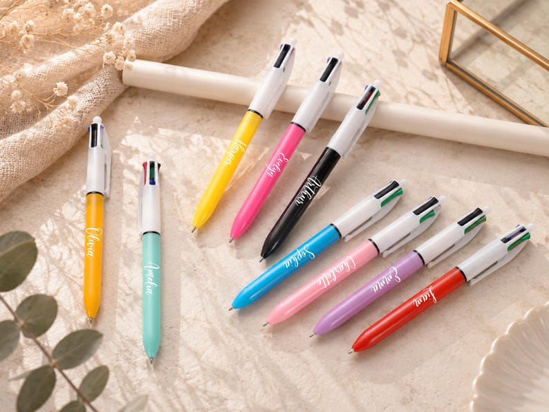 May include: A collection of colorful multi-pen ballpoint pens. Each pen has a white barrel and a different colored body, including yellow, pink, black, blue, pink, purple, and red. The pens are personalized with names in cursive script.