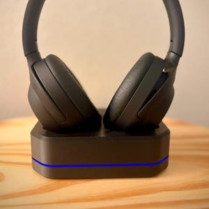May include: Black over-ear headphones resting on a black charging base. The headphones have large earcups and a padded headband. The base has a blue light strip along the front. The headphones and base are on a light brown wooden surface.