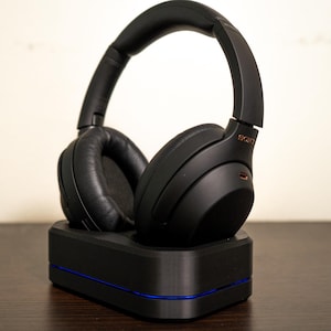 May include: Black over-ear headphones with a padded headband and earcups, resting on a black charging base with a blue accent. The headphones have the brand name "SONY" in gold lettering. The image is taken indoors.