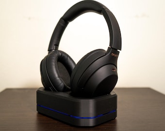 May include: Black over-ear headphones with a padded headband and earcups, resting on a black charging base with a blue accent. The headphones have the brand name "SONY" in gold lettering. The image is taken indoors.