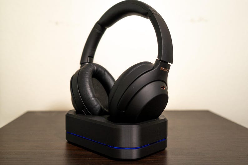 May include: Black over-ear headphones with a padded headband and earcups, resting on a black charging base with a blue accent. The headphones have the brand name "SONY" in gold lettering. The image is taken indoors.