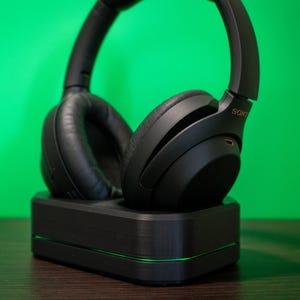May include: Black over-ear headphones resting on a black charging base with a green accent light. The headphones have a padded ear cup design and the brand name "SONY" is visible on the side.