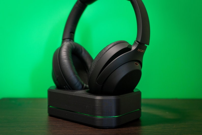 May include: Black over-ear headphones resting on a black charging base with a green accent light. The headphones have a padded ear cup design and the brand name "SONY" is visible on the side.
