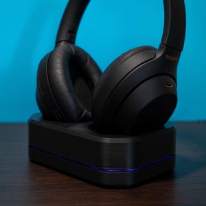 May include: Black over-ear headphones resting on a black charging base with a purple accent line. The headphones have large, cushioned earcups and a headband. The background is a solid teal color.