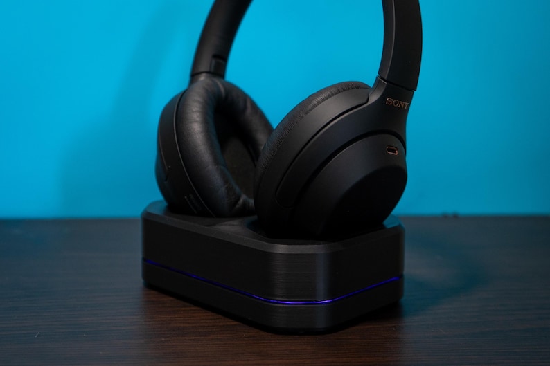 May include: Black over-ear headphones resting on a black charging base with a purple accent line. The headphones have large, cushioned earcups and a headband. The background is a solid teal color.
