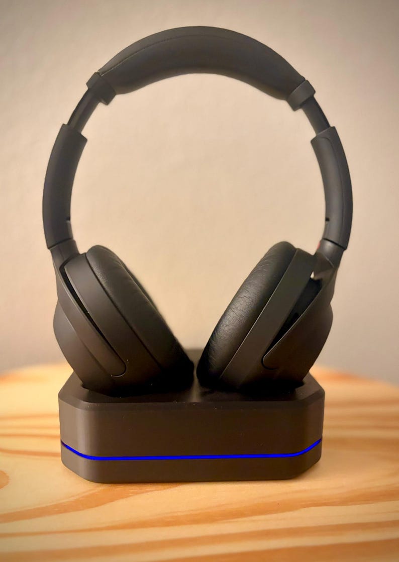 May include: Black over-ear headphones resting on a black charging base. The headphones have large earcups and a padded headband. The base has a blue light strip along the front. The headphones and base are on a light brown wooden surface.