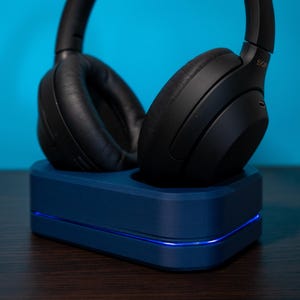 May include: Black over-ear headphones resting on a dark blue charging stand. The stand has a glowing blue light along the bottom edge. The headphones are made of black leather and plastic.