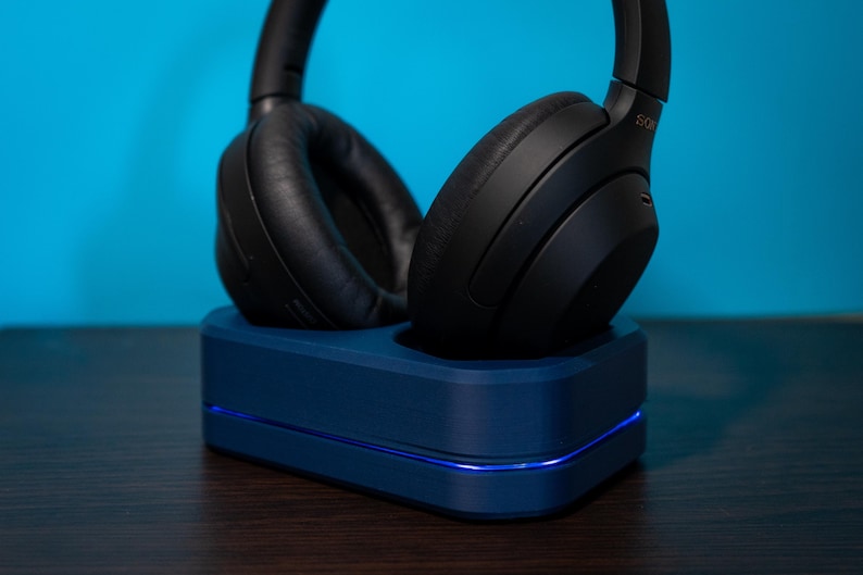 May include: Black over-ear headphones resting on a dark blue charging stand. The stand has a glowing blue light along the bottom edge. The headphones are made of black leather and plastic.