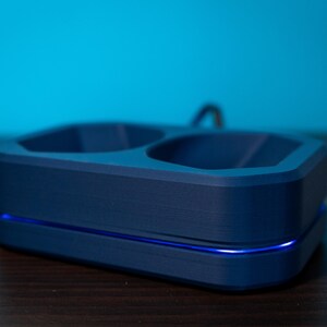 May include: A navy blue charging dock with two recessed compartments and a curved handle. The base has a blue LED light strip. The dock is on a dark wooden surface with a blue background.
