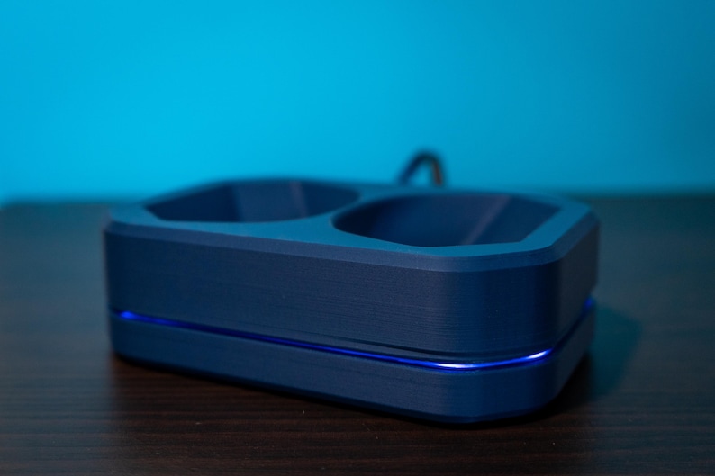 May include: A navy blue charging dock with two recessed compartments and a curved handle. The base has a blue LED light strip. The dock is on a dark wooden surface with a blue background.
