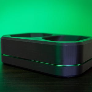 May include: A black, rectangular charging dock with two circular openings. The dock has a sleek, modern design with a thin, illuminated green line separating the top and bottom sections. The background is a gradient of green.