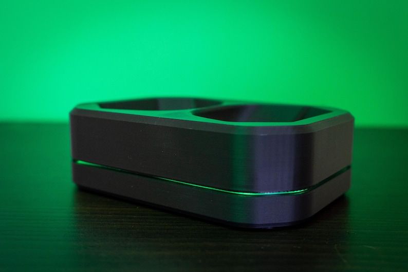May include: A black, rectangular charging dock with two circular openings. The dock has a sleek, modern design with a thin, illuminated green line separating the top and bottom sections. The background is a gradient of green.