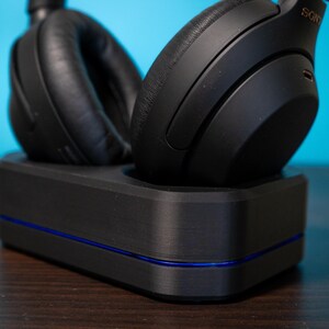 May include: Black over-ear headphones resting on a black charging dock with a blue accent line. The headphones are labeled "SONY" and have large, cushioned earcups. The dock is rectangular and sits on a dark wooden surface against a blue background.